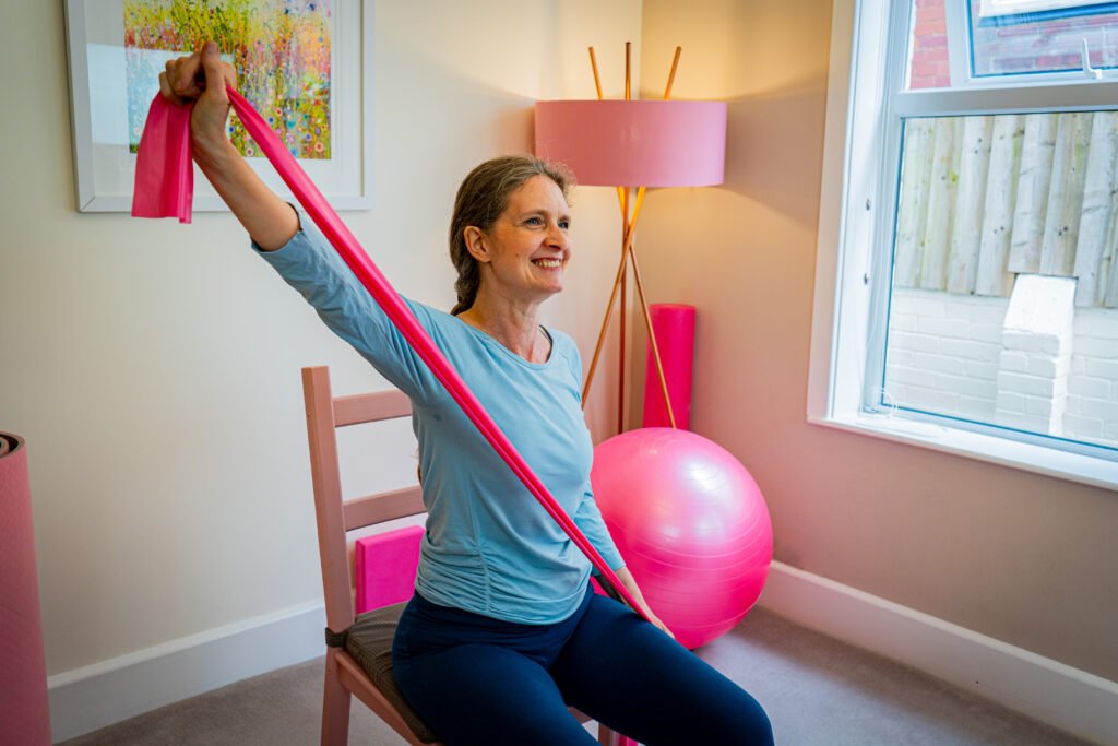 Kerry the mobilates pilates instructor sitting on a chair and using a pink exercise band to stretch her arms diagonally