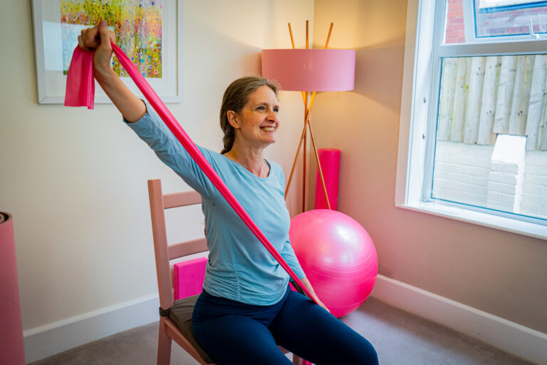 Kerry the mobilates pilates instructor sitting on a chair and using a pink exercise band to stretch her arms diagonally