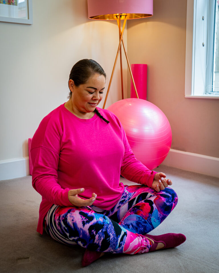 leanne English from mobilates wearing a bright pink jumper,  sitting cross legged meditating