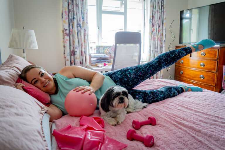 Leanne lying on her side on her bed with her leg raised, resting a hand on a pink ball. Stanley her dog, pink rsistance band and pink weights are in front of her. She is wearing blue leggings and short sleeved top.