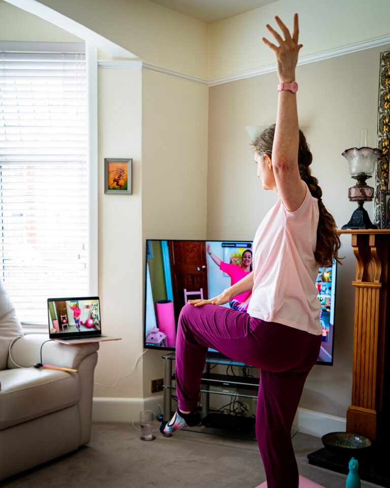 Kerry wearing a pale pink top and deep pink leggings. She is standing on her right leg with a soft knee, the left leg is lifted and she is tapping the left knee with her right hand whilst the left hand reaches up to the ceiling.
