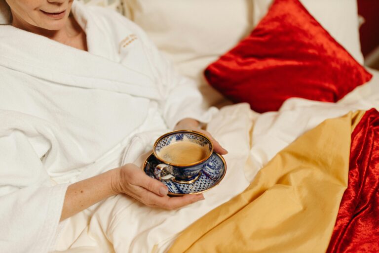 An image of a woman in bed, looking cosy, with a cup of tea.  She wearing a white dressing gown, and is wrapped up in red velvet and gold covers and cushions.