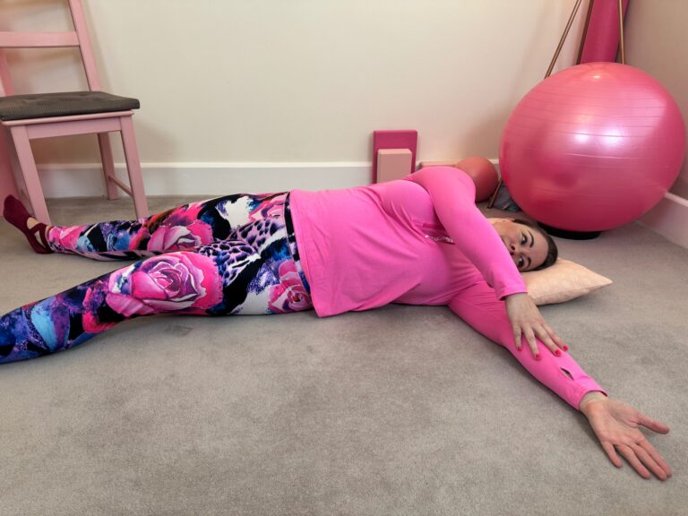 Leanne wearing a long slevve pink top and multicoloured leggings. Lying in a star position and reaching one arm across the chest.