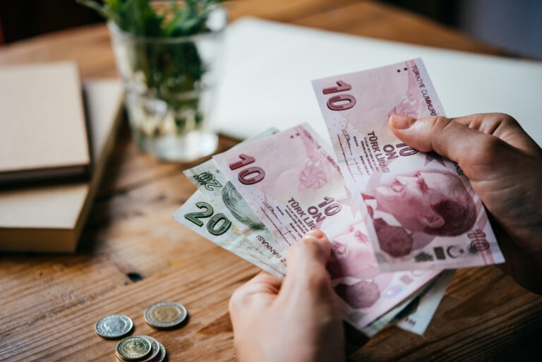 Image of 2 hands holding paper money, counting it. There is a desk, vase and some notebooks in the background.