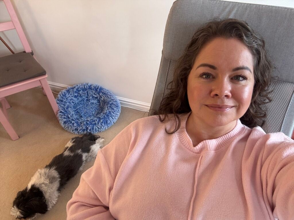 Leanne is sat in a pink sweater sits comfortably on a chair, smiling softly at the camera. Beside her, a small black and white dog is stretched out on the carpet in front of a fluffy blue pet bed, creating a cozy and serene scene at home.