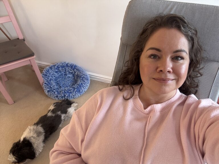 Leanne is sat in a pink sweater sits comfortably on a chair, smiling softly at the camera. Beside her, a small black and white dog is stretched out on the carpet in front of a fluffy blue pet bed, creating a cozy and serene scene at home.