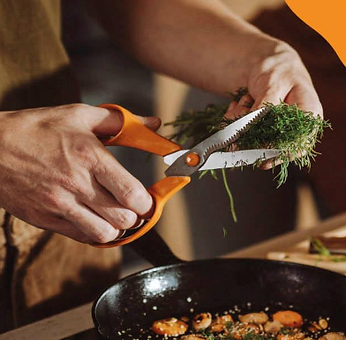 Two hands using orange handled scissors to cut herbs