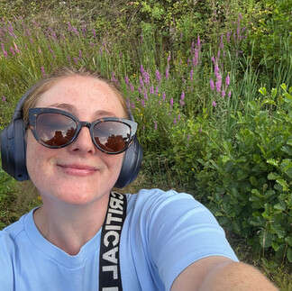 Chloe taking a selfie with greenery and purple heather behind her. She is smiling. She wears a blue tshirt, sunglasses, earphones and a bag strap across her shoulder.