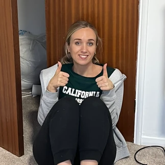 Martha sitting on the floor wearing a black outfit of black tshirt, trousers, white socks and a grey hoody. She has her thumbs up to camera and is smiling.
