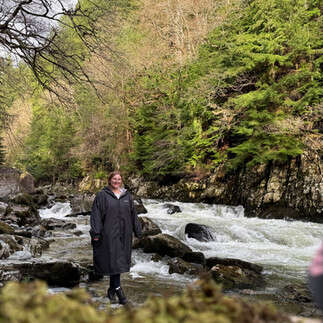 Chloe in a large black coat, smiling at the camera from a distance. She is walking next to a river. There is a big hill behind her. Lots of greenery.