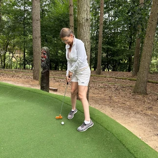 Suzanne playing mini golf, standing an dlooking happy. She is wearing light coloured shorts and a zip up light coloured top. She is about to hit the ball with her golf club.