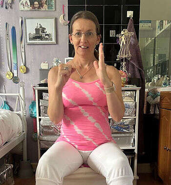 An image of our member and teacher Suzanne in a class. She is wearing a pink strappy top and white trousers. She is holding one hand in a fist and the other hand straight demonstrating a move. She is sitting and has a concentrating expression on her face.