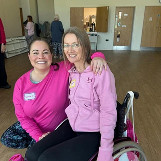 Leanne has her arm around Suzanne and is crouching next to Suzanne's wheelchair which is gorgeou sas it has pink flowers on the wheels. Both are wearing pink tops and smiling at the camera.