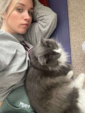 A picture of Martha taking a selfie with her cat. They are both lying on her purple Pilates mat. Martha is dressed in a grey hoody. her cat is fluffy, grey and white.