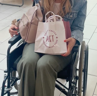 Martha sitting in a wheelchair with a bag on her lap. She has long blond hair and is smiling at the camera. She is wearing a denim jacket and green trousers.