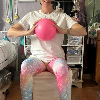 Suzanne squeezing a pink ball whilst sitting. She is wearing rainbow coloured leggings and a white tshirt and smiling.
