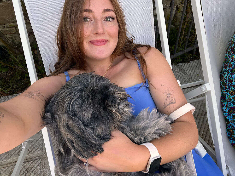 Chloe, one of our members sat on a white deck chair with a fluffy grey dong on her lap. She is wearing a blue dress and smiling at the camera, looking relaxed and happy.